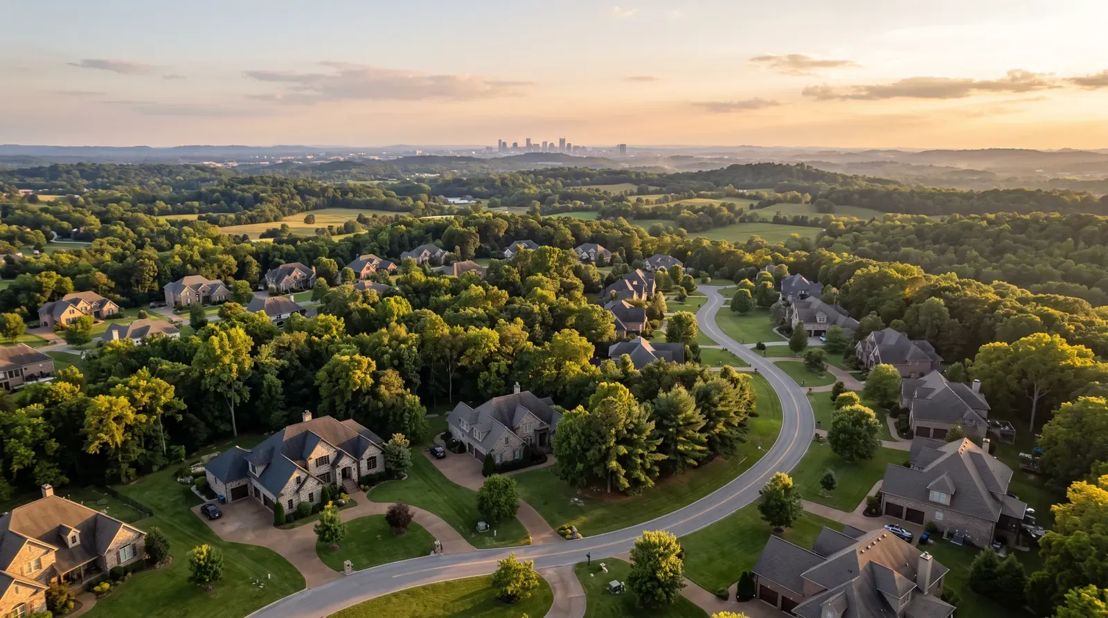 An aerial view over a green Middle Tennessee neighborhood at golden hour showing brick and stone homes, mature trees and rolling hills, a winding road and a glimpse of a city skyline far away