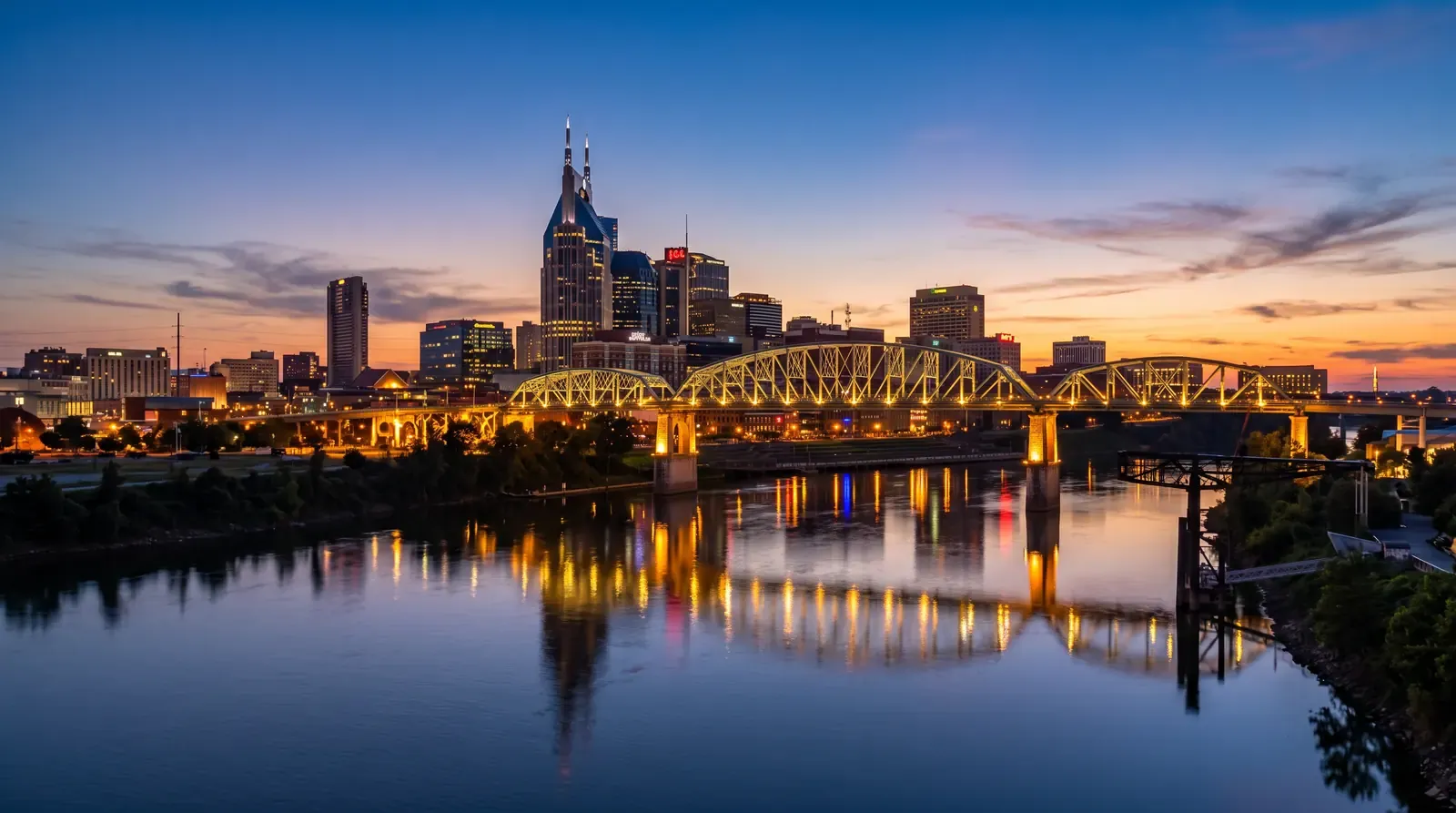 The Nashville skyline glowing at blue-hour dusk beyond the Cumberland River with a calm reflection, a warm sunset gradient in the sky and the lit pedestrian bridge